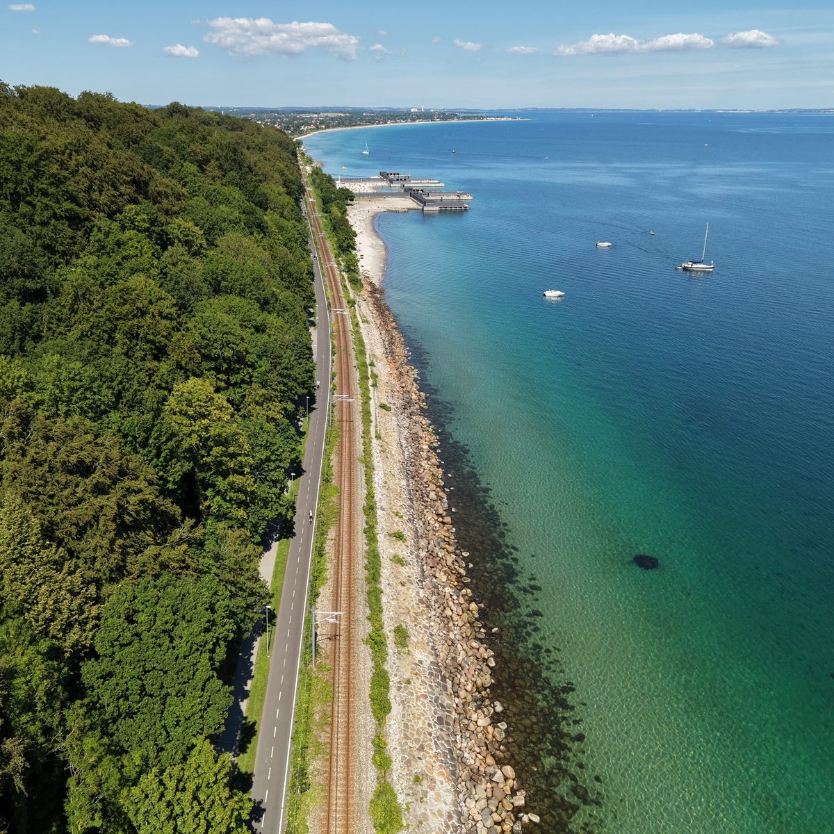 Aerial view of the forested coastline at Aarhus, with the coast road, railway line and clear turquoise shallows